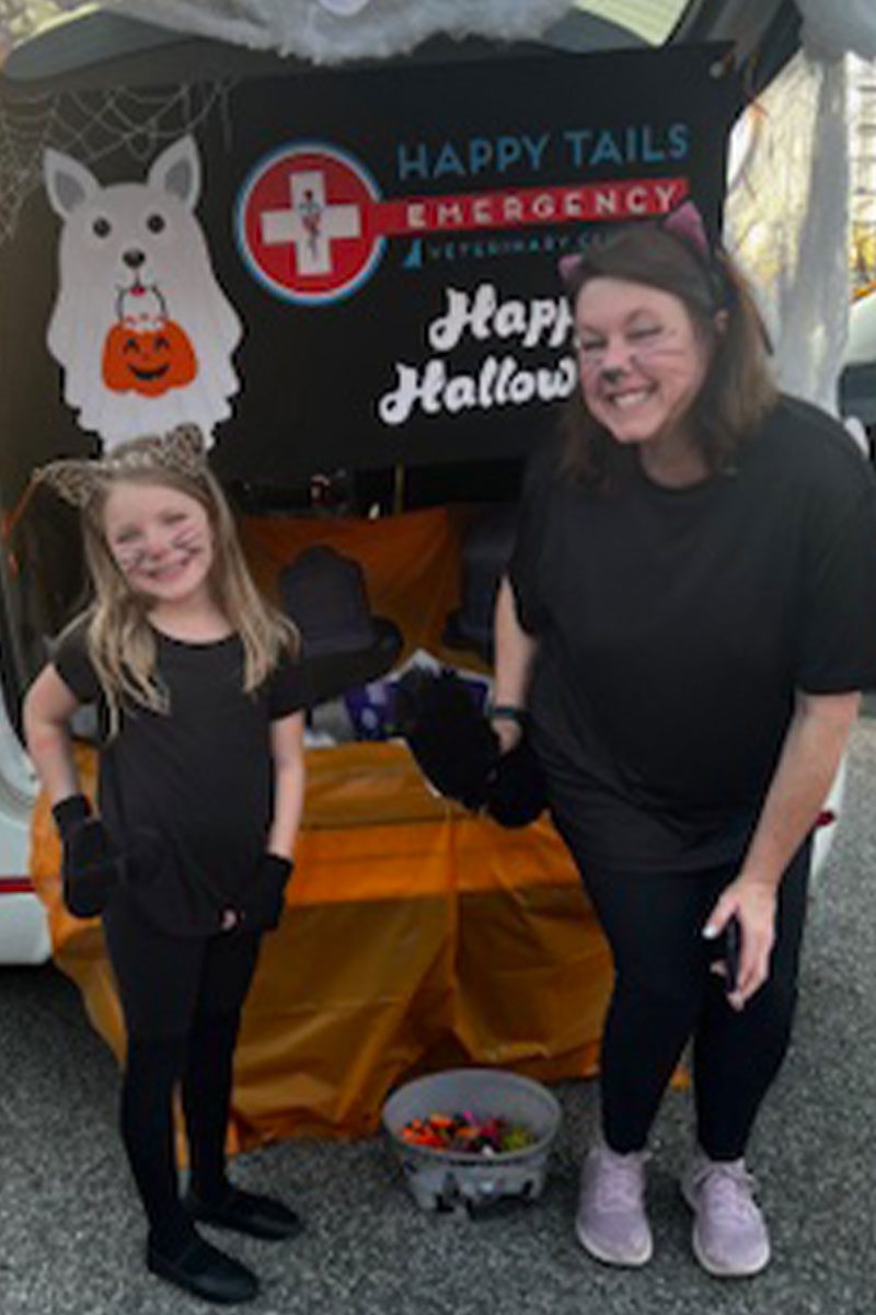 female staff member and daughter dressed up as cats smiling in front of Happy Tails trunk-or-treating car